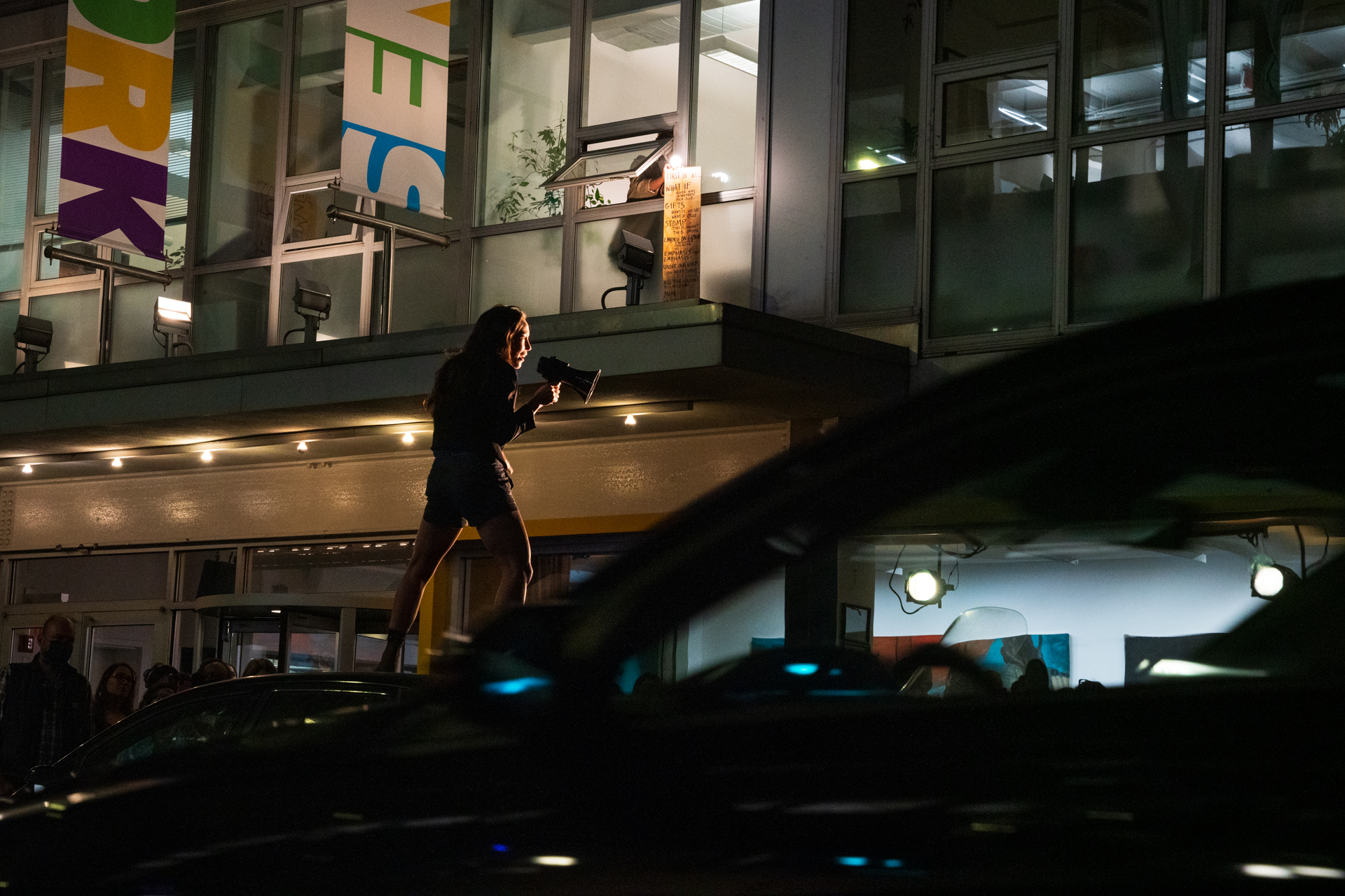 Emily Johnson stands on car , speaking through a megaphone. She is in silhouette outside New York Live Arts addressing a crowd.